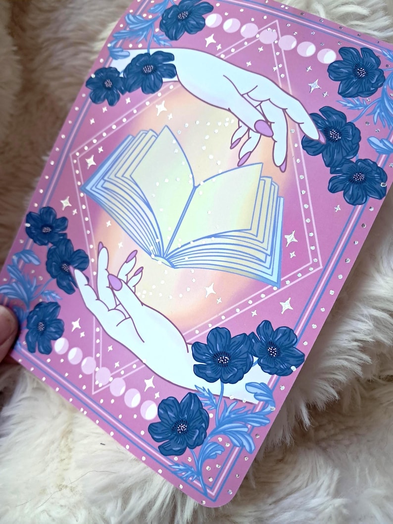 Puede incluir: Ilustraci&oacute;n rosa y azul con un libro abierto entre dos manos. La obra est&aacute; enmarcada con flores azules y fases lunares. Las manos tienen u&ntilde;as rosa claro. El fondo tiene un degradado de amarillo a rosa.
