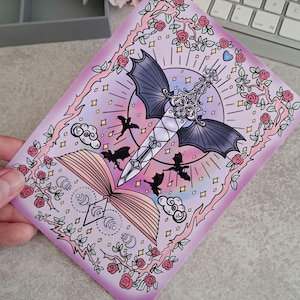 Könnte beinhalten: Ein rechteckiger Druck mit Fantasy-Thema. Das Kunstwerk zeigt ein Schwert mit Fledermausflügeln, ein offenes Buch, Fledermäuse und Rosen. Die Farbpalette umfasst Rosa, Lila und Schwarz mit einem Hauch von Weiß und Gold. Der Druck wird von einer Hand gehalten.