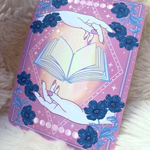 Puede incluir: Una impresi&oacute;n rectangular rosa con un libro abierto entre dos manos. Las manos son de color azul claro con u&ntilde;as rosas. Flores azul oscuro y hojas azul claro rodean el libro. El fondo tiene un patr&oacute;n de diamantes y fases lunares.