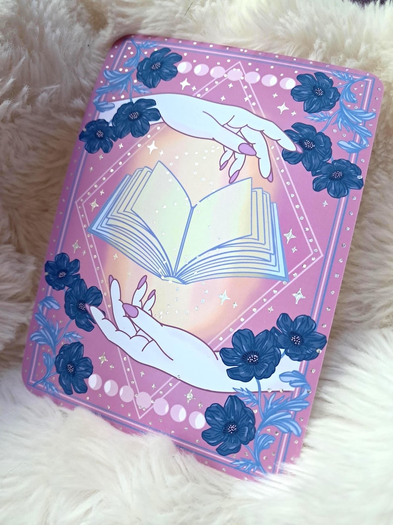 Puede incluir: Una impresi&oacute;n rectangular rosa con un libro abierto entre dos manos. Las manos son de color azul claro con u&ntilde;as rosas. Flores azul oscuro y hojas azul claro rodean el libro. El fondo tiene un patr&oacute;n de diamantes y fases lunares.