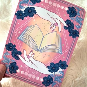 Puede incluir: Tarjeta rectangular rosa con una ilustraci&oacute;n estilizada. Dos manos blancas con u&ntilde;as rosas se extienden hacia un libro abierto. Flores y hojas azul oscuro enmarcan los bordes. El fondo tiene un dise&ntilde;o geom&eacute;trico con estrellas y fases lunares.