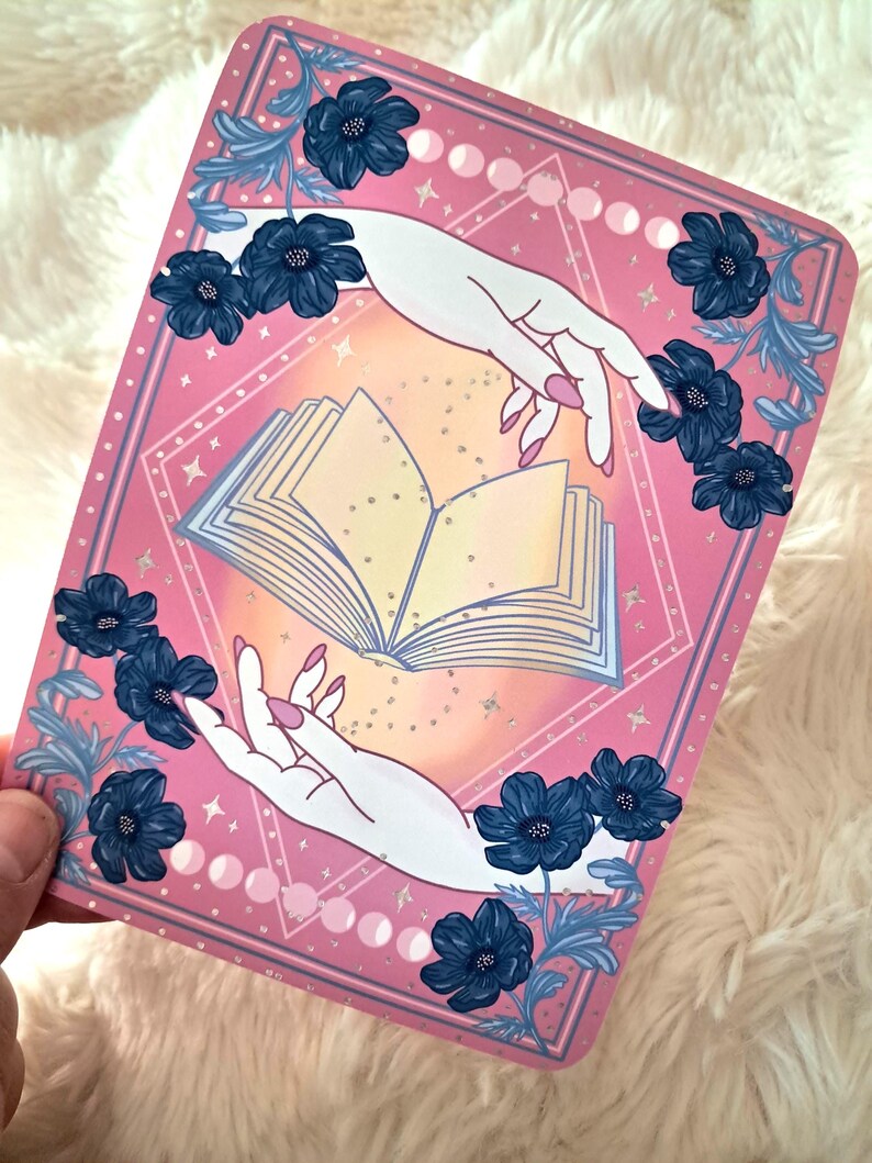 Puede incluir: Tarjeta rectangular rosa con una ilustraci&oacute;n estilizada. Dos manos blancas con u&ntilde;as rosas se extienden hacia un libro abierto. Flores y hojas azul oscuro enmarcan los bordes. El fondo tiene un dise&ntilde;o geom&eacute;trico con estrellas y fases lunares.
