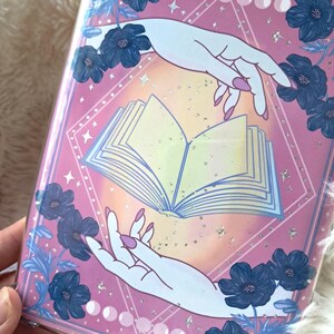 Puede incluir: Una portada de libro rosa con un dise&ntilde;o m&iacute;stico. Presenta un libro abierto, manos que se extienden hacia &eacute;l y detalles florales azules. El dise&ntilde;o incluye fases lunares y estrellas, creando una est&eacute;tica m&aacute;gica.