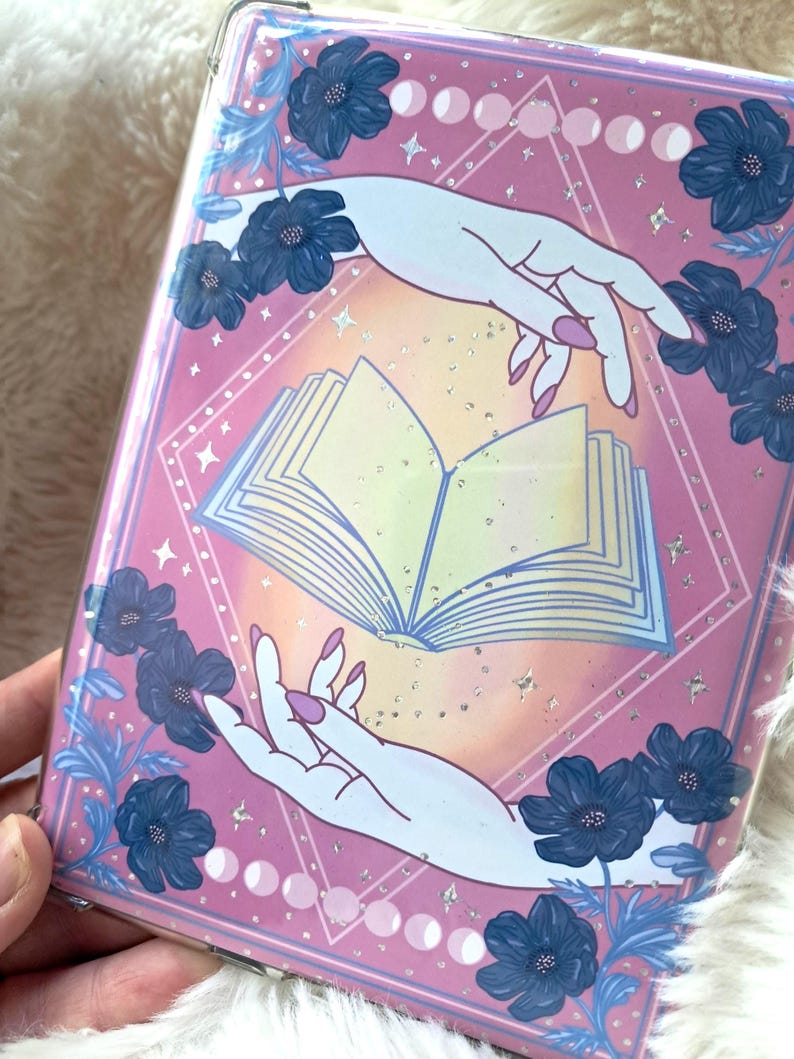Puede incluir: Una portada de libro rosa con un dise&ntilde;o m&iacute;stico. Presenta un libro abierto, manos que se extienden hacia &eacute;l y detalles florales azules. El dise&ntilde;o incluye fases lunares y estrellas, creando una est&eacute;tica m&aacute;gica.