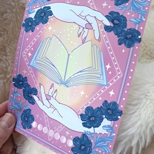 Puede incluir: Una impresi&oacute;n rectangular rosa con un libro abierto entre dos manos. Las manos son de color azul claro con u&ntilde;as rosas. Flores y hojas azul oscuro rodean el libro. La impresi&oacute;n tiene un borde en forma de diamante con estrellas y fases lunares.