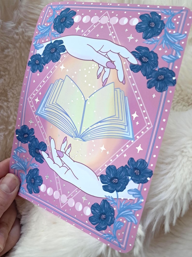 Puede incluir: Una impresi&oacute;n rectangular rosa con un libro abierto entre dos manos. Las manos son de color azul claro con u&ntilde;as rosas. Flores y hojas azul oscuro rodean el libro. La impresi&oacute;n tiene un borde en forma de diamante con estrellas y fases lunares.