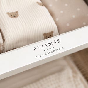 Op de afbeelding: Een wit lade-etiket met de zwarte tekst PYJAMAS BABY ESSENTIALS, geplaatst voor opgevouwen babykleding, waaronder een beige vichy-ruitstof met bloemendetails en een crèmekleurige stof met een herhalend patroon van bruine berengezichten.