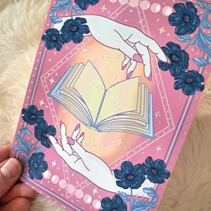 Puede incluir: Impresi&oacute;n art&iacute;stica rosa y azul con dos manos que se extienden hacia un libro abierto. La impresi&oacute;n est&aacute; decorada con flores azules, fases lunares y estrellas. Las manos tienen u&ntilde;as rosas. El tema general es m&iacute;stico y m&aacute;gico.