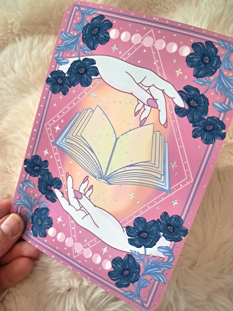 Puede incluir: Impresi&oacute;n art&iacute;stica rosa y azul con dos manos que se extienden hacia un libro abierto. La impresi&oacute;n est&aacute; decorada con flores azules, fases lunares y estrellas. Las manos tienen u&ntilde;as rosas. El tema general es m&iacute;stico y m&aacute;gico.
