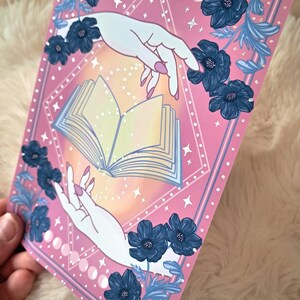 Puede incluir: Una impresi&oacute;n art&iacute;stica rosa con dos manos que se extienden hacia un libro abierto. La impresi&oacute;n est&aacute; decorada con flores azules y fases lunares. Las manos tienen esmalte de u&ntilde;as rosa. El fondo tiene un degradado de amarillo y naranja.
