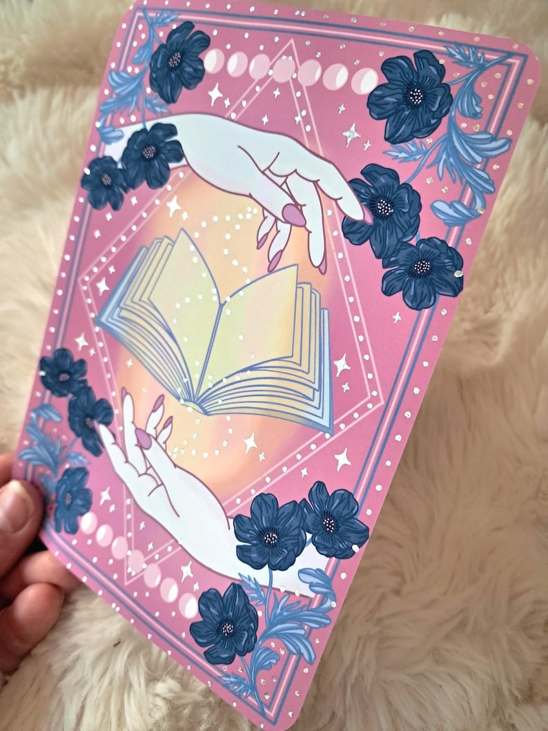 Puede incluir: Una impresi&oacute;n art&iacute;stica rosa con dos manos que se extienden hacia un libro abierto. La impresi&oacute;n est&aacute; decorada con flores azules y fases lunares. Las manos tienen esmalte de u&ntilde;as rosa. El fondo tiene un degradado de amarillo y naranja.