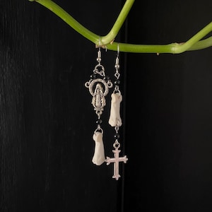May include: Silver-toned earrings with a religious theme. One earring features a pendant with a Virgin Mary design and black beads. The other earring has a bone-shaped charm and a silver cross pendant. The earrings hang from a green vine against a black background.