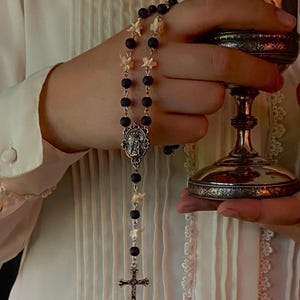 May include: A rosary with black beads and bone-shaped accents, featuring a silver cross and a pendant with a religious figure. A silver chalice is held in the background.