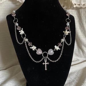 Könnte beinhalten: Eine silberfarbene Halskette mit einem Kreuzanhänger, rosa herzförmigen Perlen und weißen floralen Akzenten. Die Halskette hat mehrere Stränge aus feiner Kette und kleinen, runden Perlen. Sie wird auf einer schwarzen Schaufensterpuppe präsentiert.