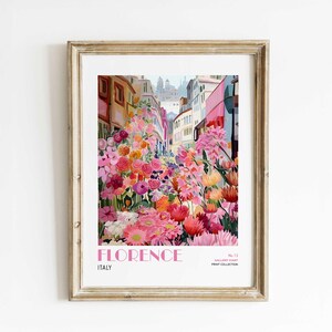 Puede incluir: Una impresión artística enmarcada que presenta una vibrante escena callejera en Florencia, Italia, llena de flores coloridas. La obra de arte muestra una variedad de flores rosas, naranjas y moradas que bordean la calle, con edificios al fondo. La palabra "FLORENCE" está impresa en rosa.