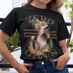 以下が含まれることがあります： 花と緑に囲まれたキツネのグラフィックがプリントされた黒いTシャツ。Tシャツには茶色の文字で「ZERO GIVEN」とプリントされています。