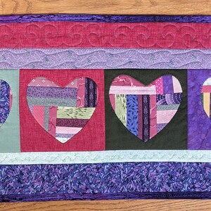 May include: A quilted table runner with a purple, green, and pink color scheme. Four hearts are cut out of the fabric and are filled with a patchwork of different fabrics. The hearts are arranged in a row in the center of the runner.