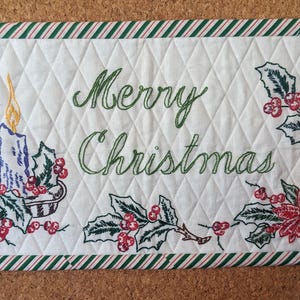 May include: A rectangular quilted wall hanging with a white background and a red and green striped border. The center features the embroidered text Merry Christmas in green, surrounded by embroidered holly, berries, and a lit candle.