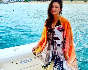 May include: A woman wearing a long, orange and white patterned dress with a large print of a woman's face on the front. The dress has a V-neck and long sleeves. The woman is standing on a boat with the ocean in the background. The boat has the text "Blue Sea" and "www.blueskycabo.com" on the side.