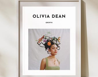 May include: Framed art print with the text "OLIVIA DEAN" and "GROWTH." The print depicts a person adorned with a floral headpiece and a green top. The artwork is displayed on a white wall with wooden flooring.