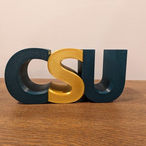 May include: A three-dimensional, block-style sign spelling out "CSU" in gold and teal blue.