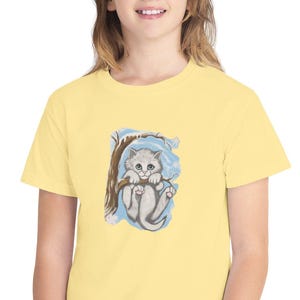 Könnte beinhalten: Ein hellgelbes T-Shirt mit einer Aquarellillustration eines grauen Kätzchens auf einem Ast. Das Kätzchen hat blaue Augen und ist vor einem blau-weißen Hintergrund platziert. Das T-Shirt ist aus einem weichen Material.