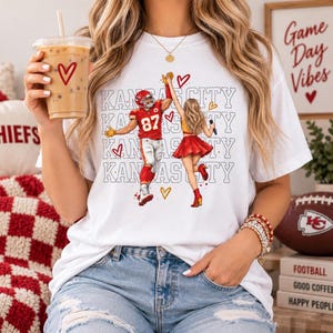 May include: A white short-sleeved t-shirt featuring a graphic of a football player in a red jersey with the number 87 and a cheerleader in a red skirt high-fiving. The text Kansas City is repeated in the background with small red heart icons.