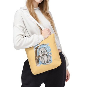 Peut inclure: Un sac fourre-tout jaune clair avec une illustration à l'aquarelle d'un chaton gris accroché à une branche d'arbre. Le chaton a les yeux bleus et se détache sur un fond bleu clair. Le sac a une sangle blanche.