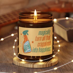 May include: A lit candle in a brown glass jar with a label that reads "magically turn all that crappiness into happiness." The label also features a cartoon image of a spray bottle. The candle sits on a wooden coaster, surrounded by fairy lights.