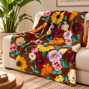 Op de afbeelding: Een bruine plaid met een levendig bloemenpatroon van gele zonnebloemen, donkerrode, roze, oranje en lichtpaarse bloesems, samen met groene bladeren en stengels, gedrapeerd over de armleuning en zitting van een lichtgekleurde bank.