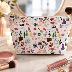 May include: A white zipped pouch featuring a repeating pattern of colourful icons including guitars, crowns, hearts, disco balls, cowboy boots, pianos, the number 13, planets, butterflies, feathers, and musical notes printed across the fabric surface.