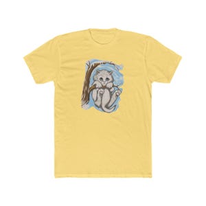 Pode incluir: Camiseta amarela clara com uma ilustração em aquarela de um gatinho cinza em um galho de árvore. O gatinho tem olhos azuis e está em um fundo azul claro. A camiseta é feita de um material macio.