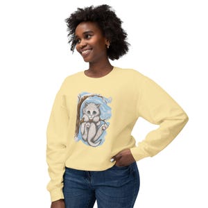 May include: A light yellow sweatshirt featuring a whimsical illustration of a gray kitten clinging to a tree branch. The artwork includes blue accents and a snowy background. The long-sleeved top is made of a soft material.