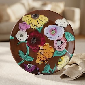 May include: A round ceramic plate with a glossy brown background featuring a vibrant floral pattern. The design includes large yellow, deep red, light purple, pink, and white flowers with green leaves scattered across the surface.