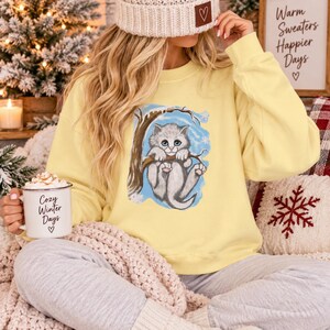 May include: A pale yellow crewneck sweatshirt featuring a graphic illustration of a fluffy gray kitten clinging to a brown tree branch against a light blue background. The kitten has large blue eyes and is surrounded by white snowflakes.