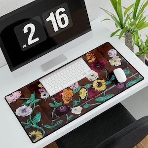 May include: A rectangular brown desk mat with a floral design featuring sunflowers, roses, and other flowers in shades of yellow, pink, purple, and white. A white keyboard and mouse sit on the mat. The mat is on a white desk with a computer monitor.
