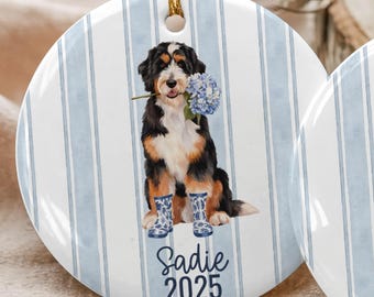 May include: Two round ceramic ornaments with gold string hangers. One features a black, brown, and white dog holding blue flowers, wearing blue boots, and the name "Sadie 2025". The other shows a golden retriever with blue flowers, blue boots, and the name "Duke 2025".