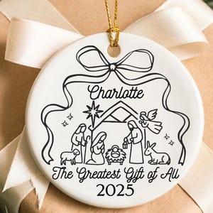 May include: A white ceramic Christmas ornament with a gold string, featuring a black and white nativity scene design. The ornament is personalized with the name "Charlotte" and the text "The Greatest Gift of All 2025". It rests on a wrapped gift.