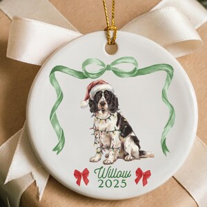 May include: A white ceramic Christmas ornament featuring a watercolor-style Springer Spaniel wearing a Santa hat and string lights. The ornament has green ribbon accents, red bows, and the name "Willow 2025". The ornament hangs from a gold string.