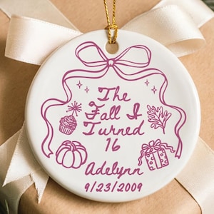 May include: A white ceramic ornament with a gold string, featuring the text "The Fall I Turned 16 Adelynn 9/23/2009" in purple. The ornament is decorated with a bow, stars, a cupcake, a pumpkin, and a gift. It rests on a wrapped gift with a white ribbon.
