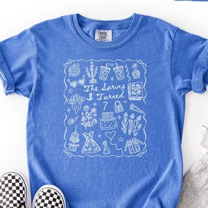 May include: A blue t-shirt with the text "The Spring I Turned 7" and a white doodle design. The shirt is paired with gray sweatpants and black and white checkered slip-on shoes. The shirt is labeled "Flo Blue".
