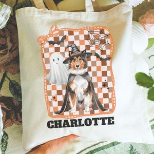 May include: Cream-colored tote bag featuring a Halloween-themed design. The design includes a dog wearing a witch hat and cape, a ghost, bats, and a spiderweb against a checkered background. The name "CHARLOTTE" is printed at the bottom.