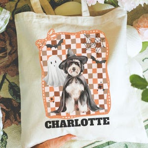 May include: A cream-colored tote bag featuring a Halloween-themed design. The design includes a dog wearing a witch hat and cape, a ghost, bats, and a spiderweb on a checkered background. The name "CHARLOTTE" is printed at the bottom.