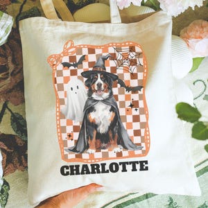 May include: A cream-colored tote bag featuring a Halloween-themed graphic. The design includes a dog wearing a witch hat and cape, a ghost, bats, and a spiderweb, set against a checkered background. The name "CHARLOTTE" is printed below the graphic.