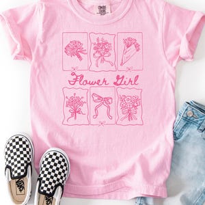 May include: A light pink t-shirt with a floral design and the words "Flower Girl". The shirt is paired with denim shorts and black and white checkered slip-on shoes. The text "Blossom" and "Comfort Colors" are also visible.