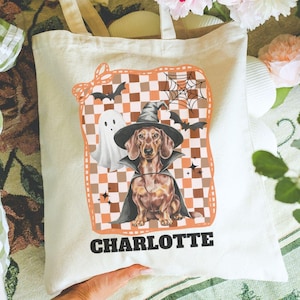 May include: Cream-colored tote bag featuring a Halloween-themed dachshund illustration. The design includes a dachshund wearing a witch hat and cape, a ghost, bats, and a checkered pattern. The name "CHARLOTTE" is printed at the bottom.