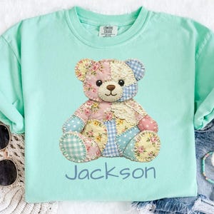 May include: A pale turquoise t-shirt with a patchwork teddy bear design in floral and gingham patterns. The name "Jackson" is printed in blue script below the bear. The shirt is part of the "Island Reef" collection.