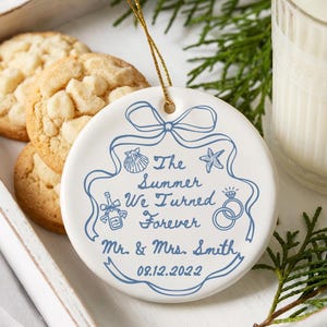 May include: A white ceramic ornament with blue text and a gold string. The text reads "The Summer We Turned Forever" with a date and names. The ornament is on a white tray with cookies and a glass of milk.