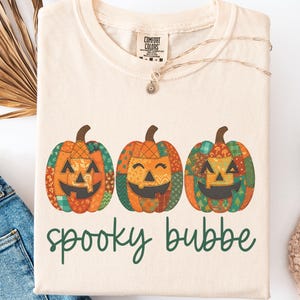 May include: Cream-coloured t-shirt with three smiling pumpkin designs in orange, green, and brown patchwork. The words "spooky bubbe" are printed in green script below. The shirt is part of a Halloween or autumn-themed collection.