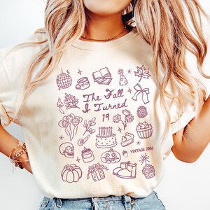 May include: Ivory-colored t-shirt with a purple graphic design. The design includes the text "The Fall I Turned 19 Vintage 2006" surrounded by various illustrations such as a skull, cake, and pumpkin.